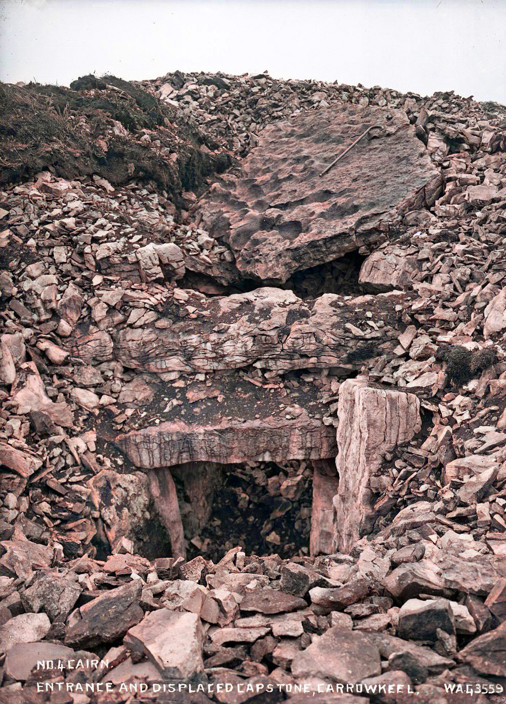 the entrance to Cairn F prior to digging in 1911. The large slab above the entrance was the capstone of the chamber, with a walking stick for scale. The workers smashed this slab because it was too heavy to lift.